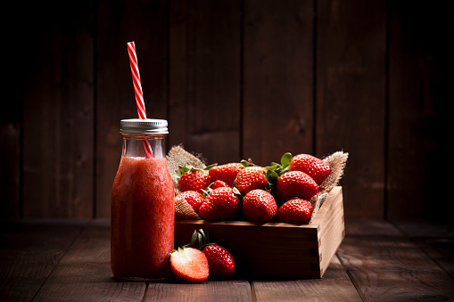 Horizontal shot of a strawberry smoothie in a glass bottle with a red and white drinking straw on rustic wood table. A small wooden crate filled with strawberries is at the right of the bottle and some fruits are out of the crate. Predominant colors are red and brown. Low key DSRL studio photo taken with Canon EOS 5D Mk II and Canon EF 70-200mm f/2.8L IS II USM Telephoto Zoom Lens