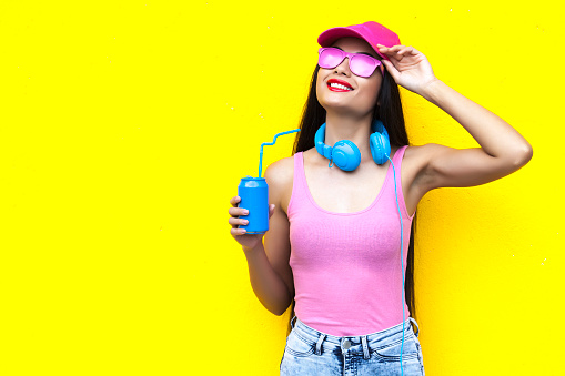 Woman wearing pink vest, blue headphones and holding blue can in front of yellow wall background