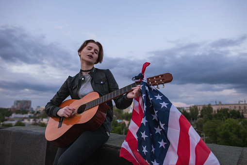 Singer with the US flag playing the guitar on cloudy sky background. Acoustic concert on the roof. A teenager on vacation at a party on Independence Day