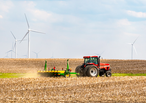 Farmer plowing land next to wind farm with wind turbines rotating in background
