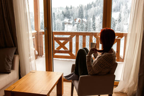 istock Woman sitting in a comfortable chair drinking coffee and looking through window at snow covered mountain. 695246796
