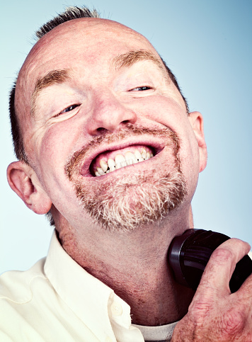 A mature, bearded man is shaving his jawline and neck with an electric razor, grimacing with clenched teeth.