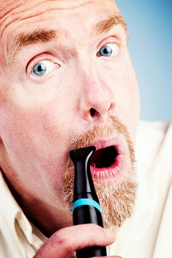 A mature, bearded man is trimming his moustache with asn attachment from his electric razor and pulling a face to keep his lip taut.