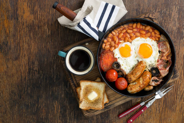 Full English breakfast with sausages, bacon, fried eggs, beans, toast and cup of coffee on wooden table with copy space Full English breakfast with sausages, bacon, fried eggs, beans, toast and cup of coffee on wooden table with copy space, top view english breakfast stock pictures, royalty-free photos & images
