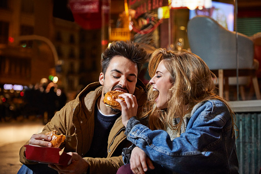 Cheerful woman feeding burger to man. Male and female friends are sitting outside restaurant. They are in casuals at night.