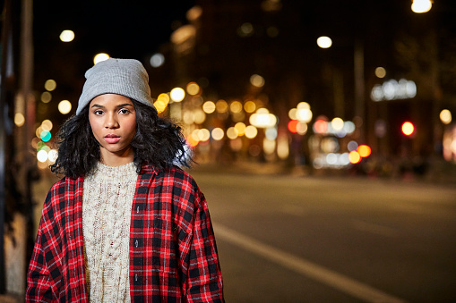 Portrait of young woman standing on roadside. Female is in illuminated city at night. She is wearing casuals.