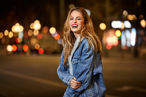 Portrait of smiling young woman standing on street. Cheerful female with brown hair wearing denim jacket. She is in city at night.