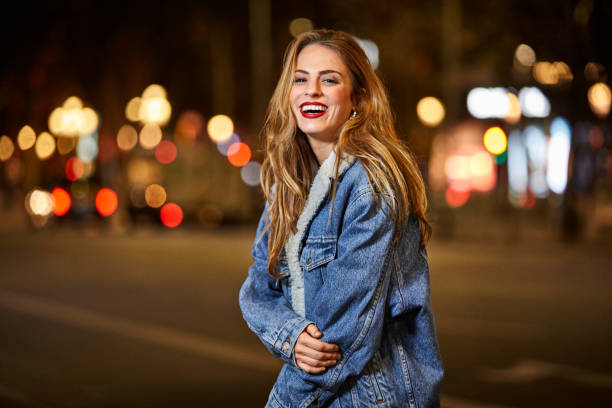 Portrait of happy woman standing on road at night Portrait of smiling young woman standing on street. Cheerful female with brown hair wearing denim jacket. She is in city at night. denim jacket stock pictures, royalty-free photos & images