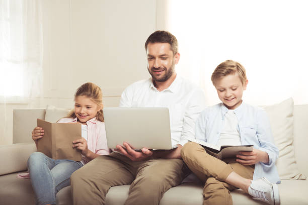 istock smiling father using laptop while his daughter and son reading books and sitting on sofa 695450886