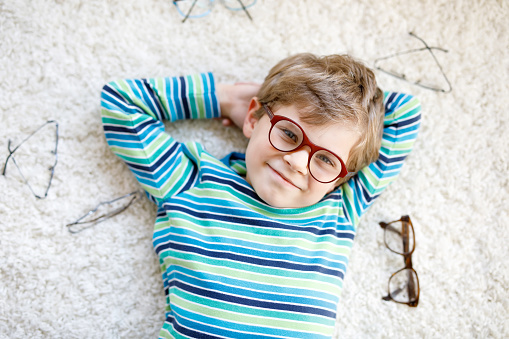 Close-up portrait of little blond kid boy with different eyeglasses on white background. Happy smiling child in casual clothes. Childhood, vision, eyewear, optician store. Boy choosing new glasses