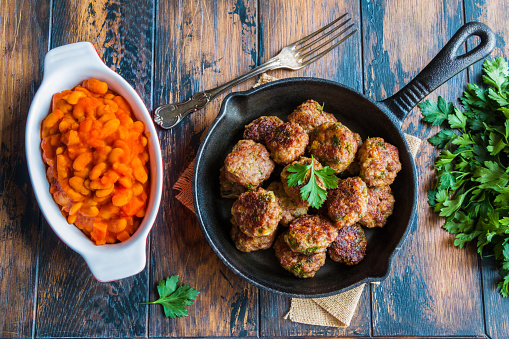 Homemade roasted beef meatballs in cast-iron skillet and beans baked in tomato sauce in baking dish on wooden table in kitchen, fresh parsley, vintage fork, top view.