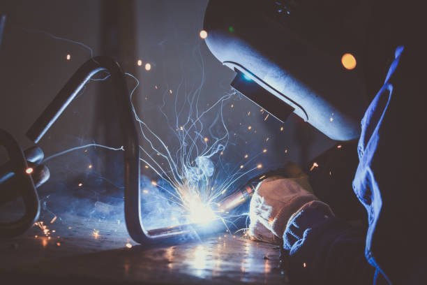 Worker with protective mask welding steel pipe on a work bench and producing  sparks