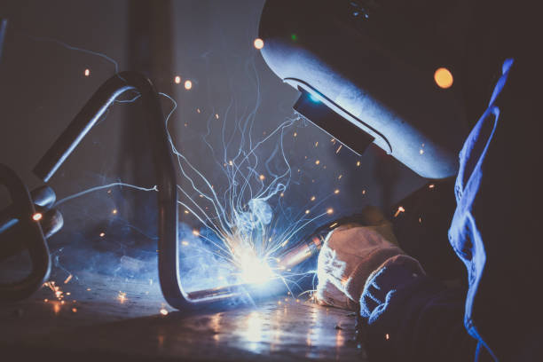 Worker with protective mask welding steel pipe on a work bench and producing  sparks stock photo