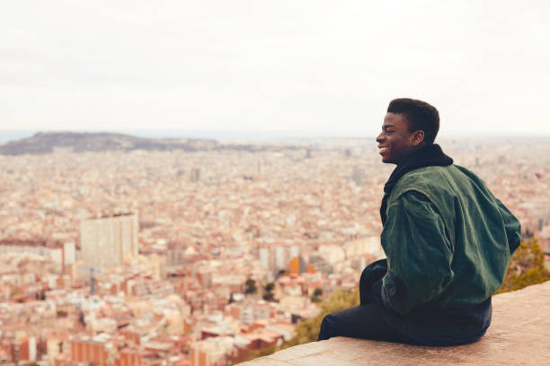 Smiling young man sitting on building terrace. Male is looking at cityscape. He is wearing casuals.