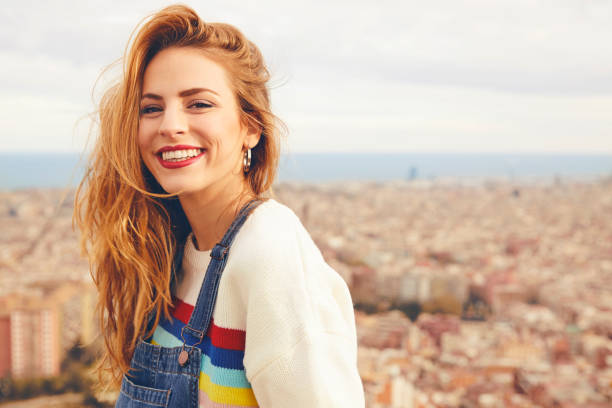 Portrait of happy young woman against cityscape Portrait of beautiful smiling woman against cityscape. Young female is wearing bib overalls. She is having leisure time. redhead photos stock pictures, royalty-free photos & images