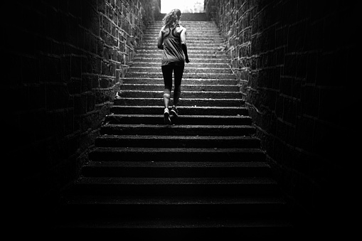 An adult woman training on a large set of outdoor steps, running and employing other stairway exercise variations.  Black and white image of her sprinting up the stairway from behind.