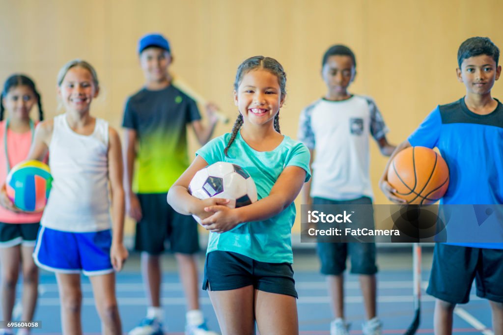 Many Activities A multi-ethnic group of kids are standing in a gymnasium. They are holding a basketball, volleyball, hockey stick, skipping rope, baseball bat, and soccer ball. They are smiling. Child Stock Photo