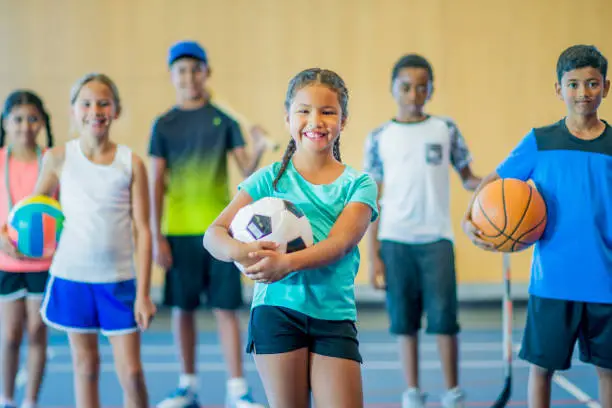 A multi-ethnic group of kids are standing in a gymnasium. They are holding a basketball, volleyball, hockey stick, skipping rope, baseball bat, and soccer ball. They are smiling.