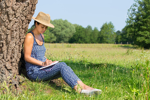 Young woman sitting against tree trunk writing in meadow. The indian woman sits in nature near a green meadow with flowers and green grass. She's wearing a straw hat on this sunny day in summer season. The person likes it to relax and enjoy the freedom and silence of a natural environment. It gives her inspiration to write in a creative way. In the background you see trees of a forest.