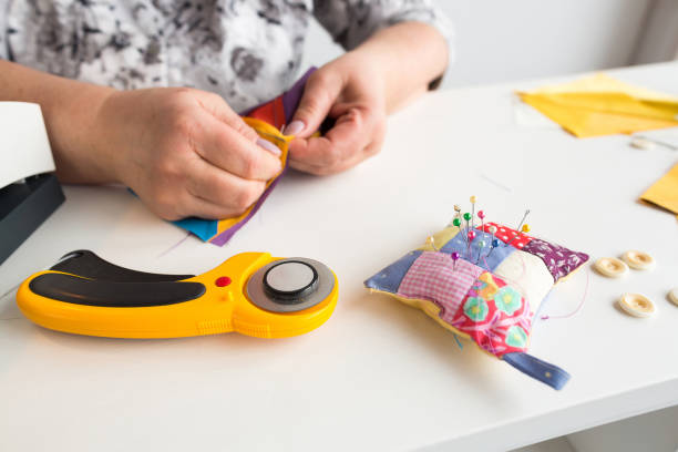 istock needlework and hand quilting - tailor hands sew multicolored patchwork fabric with the use of needles, cushions for pins, buttons and rotary fabric cutters on white background 695604546