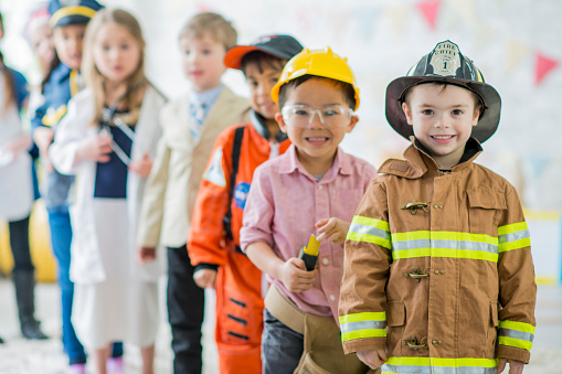 A multi-ethnic group of young children are indoors. They are wearing costumes to show what future jobs they want. A chef, construction worker, police officer, astronaut, firefighter, and doctor are posing together in a line and smiling at the camera.