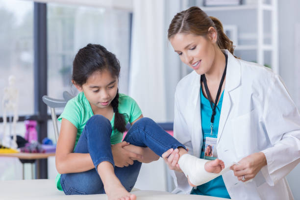 Confident physician wraps girl's injured ankle Pretty mid adult female physician wraps a young girl's injured foot. The girl is sitting on an exam table. Crutches are behind the girl. emergency medicine stock pictures, royalty-free photos & images