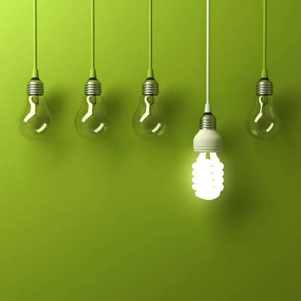 Photo of One hanging energy saving light bulb glowing different standing out from unlit incandescent bulbs with reflection on green background, leadership and different creative idea concept