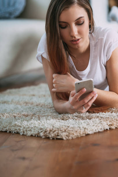 istock Young woman checking her smart phone lying on carpet 695704474