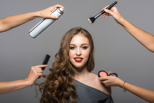 Makeup and hairstyle process, portrait of beautiful young woman and many hands with a brush, powder, comb and hairspray. Studio shot, gray background