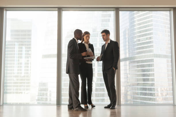 istock Three employees discussing documents standing near big window in office 695760072