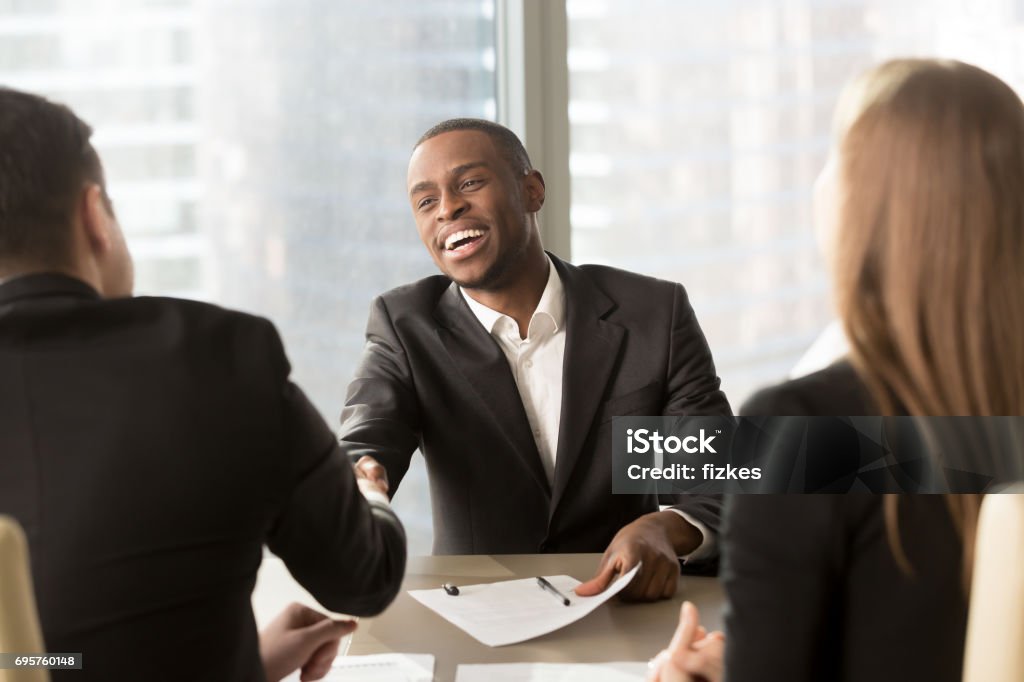 Excited smiling black businessman handshaking white partner at multiracial meeting Excited smiling black businessman handshaking white partner at meeting, successful african applicant getting hired, got a job, satisfied multiracial businessmen shaking hands after signing document Job Interview Stock Photo