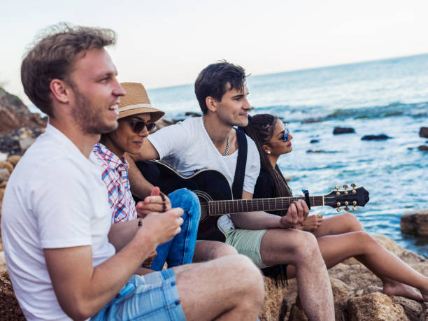 istock friends sitting on stones on beach. man is playing guitar. 695802748