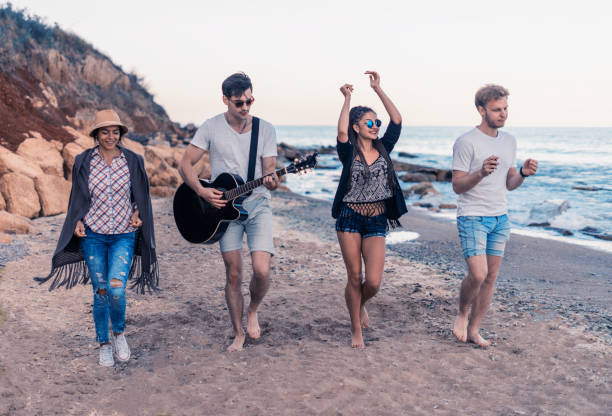 group of young and cheerful friends walking on beach, One man is playing guitar. Dancing and singing on wild beach. Happy couples