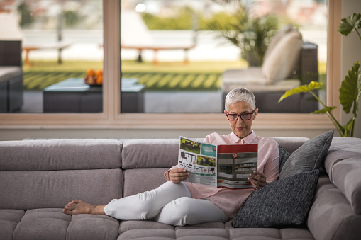 Elderly woman relaxing on the sofa in the living room and reading a magazine.