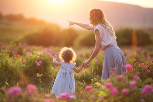 Beautiful child girl with young mother are wearing casual clothes walking in roses garden over sunset lights stock photo