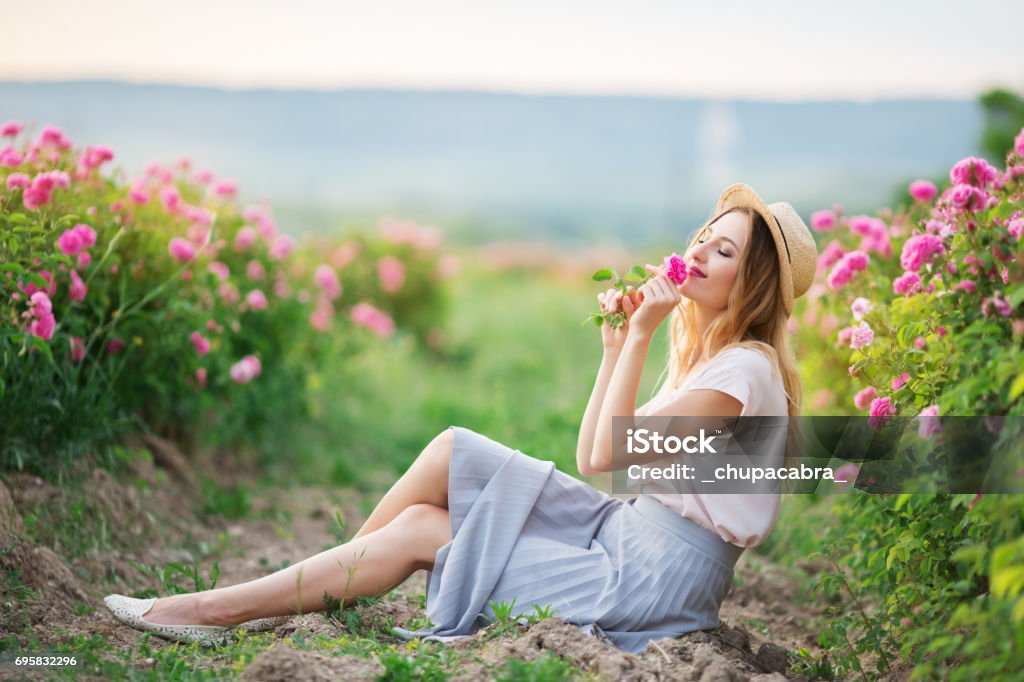 Beautiful young girl is wearing straw hat sitting in a garden with pink roses Beautiful pretty woman is sitting near roses in a garden. The concept of perfume advertising. Springtime Stock Photo