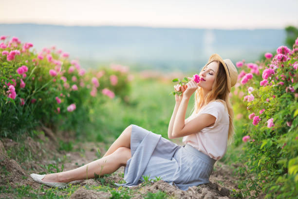 Beautiful young girl is wearing straw hat sitting in a garden with pink roses stock photo