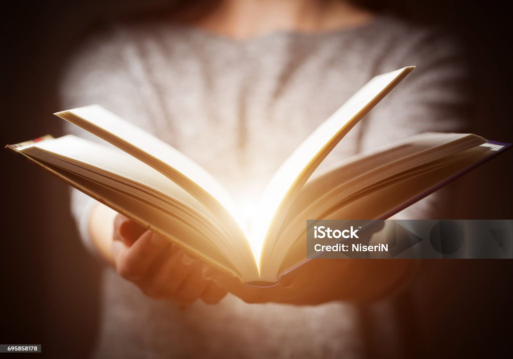 Light coming from book in woman&#39;s hands in gesture of giving Light coming from book in woman&#39;s hands in gesture of giving, offering. Concept of wisdom, religion, reading, imagination. Storytelling Stock Photo