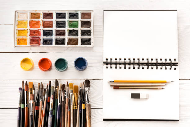 istock Top view of various paintbrushes, paints and blank open notebook on wooden table 695869278