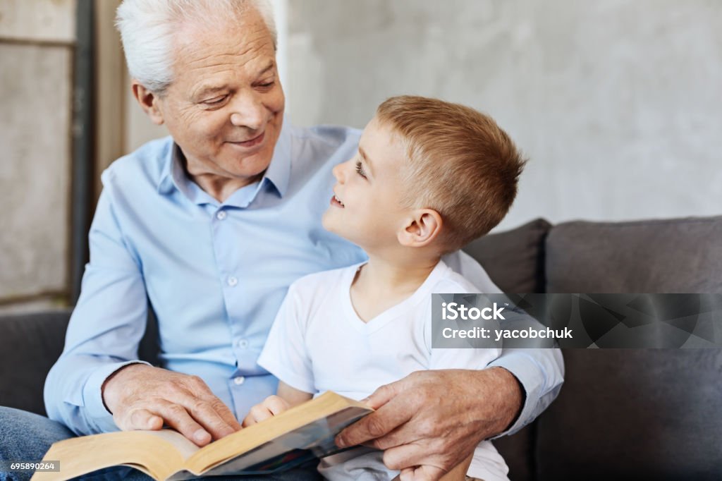 Cute energetic kid enjoying time with grandpa Strong bond. Committed bright excited grandpa and his child having some good time together while talking a lot and reading interesting books at home Grandfather Stock Photo