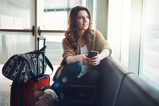 Shot of a young woman sitting in an airport with her luggage and holding her phone while looking outside