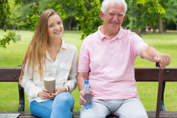 Man on a park bench accompanied by caregiver Man sitting on a park bench accompanied by young female caregiver united states senate committee on health education labor and pensions stock pictures, royalty-free photos & images