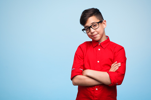 Small boy in shirt and glasses looking smart and posing with hands crossed on blue.