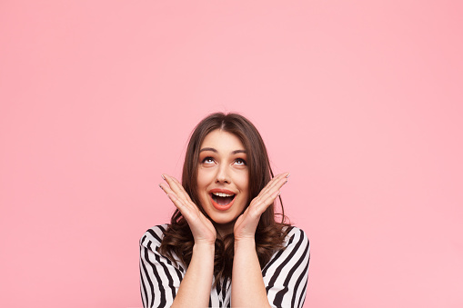 Young girl in striped shirt posing with hands on pink background looking up in excitement.
