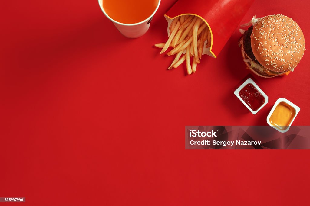 Fast food dish top view. Meat burger, potato chips and glass of drink on red background. Takeaway composition Fast food dish top view. Meat burger, potato chips and glass of drink on red background. Takeaway composition. Wrapped French fries, hamburger, mayonnaise and ketchup sauces on red desk. Copy space Fast Food Restaurant Stock Photo