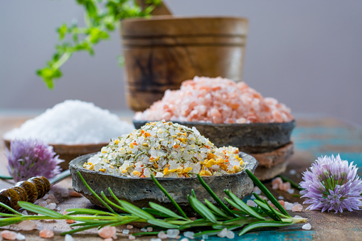 Different types of natural salt in stone bowls on wooden surface. White sea salt, pink Himalayan salt, Spiced salt with rosemary
