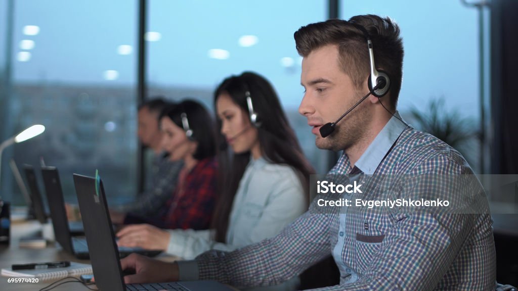 Young man in call center Young handsome men in checked shirt and headset working in call center in modern worldwide office he talking and looking on screen of desktop computer Call Center Stock Photo