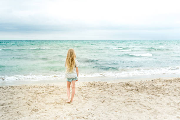 Young blonde girl standing on the beach looking out over the ocean