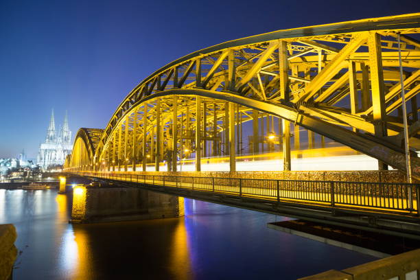 Long exposure of the Hohenzollern Bridge in Cologne that crosses the Rhine River from south to north of the city. This bridge serves trains and pedestrians and is one iconic place to visit there because of the views of the town and the river with boats, also because it has thousans of locks on one of its fences. People hang locks as display of strong love and wishes. The cathedral can be see in the background in this image.