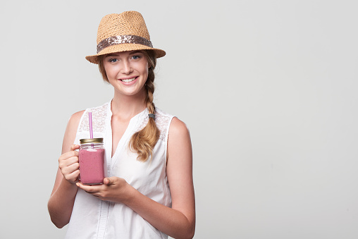 Smiling girl holding jar tumbler mug with pink smoothie drink, portrait over grey background with blank copy space
